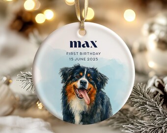 May include: A round, white ceramic ornament with a watercolor painting of a dog. The dog is black, brown, and white with its tongue out. The ornament has the name "max" and the text "FIRST BIRTHDAY 15 JUNE 2025". A gold ribbon is attached.