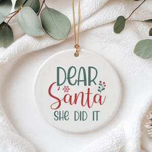 May include: A white ceramic Christmas ornament with the text "DEAR Santa SHE DID IT" in green and red script. The ornament has a gold-colored string for hanging and is decorated with a snowflake and berries.