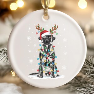 May include: A white ceramic Christmas ornament featuring a black Labrador Retriever wearing a Santa hat and reindeer antlers. The dog is wrapped in colorful Christmas lights and surrounded by snowflakes.
