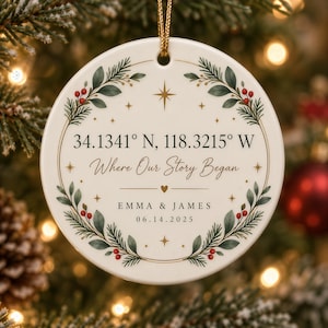 May include: A round white ceramic ornament featuring a green pine and red berry wreath design. The text reads 34.1341° N, 118.3215° W, Where Our Story Began, EMMA & JAMES, and 06.14.2025. It includes gold star accents and a gold hanging cord.