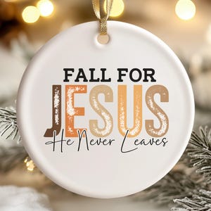 May include: White ceramic Christmas ornament with the text "FALL FOR JESUS He Never Leaves" in black, brown, and orange. The ornament has a gold ribbon for hanging and is decorated with a festive design.