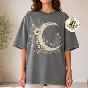 Könnte beinhalten: Ein anthrazitfarbenes Oversize-T-Shirt mit einer großen, cremefarbenen Grafik einer Mondsichel, die mit verschiedenen Wildblumen und Gänseblümchen verziert ist. Ein kreisförmiges Abzeichen oben rechts enthält den Text 100% ORGANIC VINTAGE TEE.