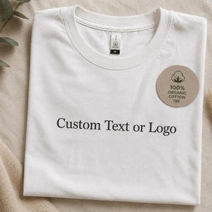 Könnte beinhalten: Ein weißes T-Shirt mit Rundhalsausschnitt und dem schwarzen Schriftzug Custom Text or Logo auf der Brust. Ein rundes, beiges Etikett ist am T-Shirt befestigt und zeigt eine Baumwollgrafik sowie den Text 100% ORGANIC COTTON TEE.
