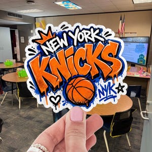 May include: A die-cut sticker featuring the text NEW YORK KNICKS in bold orange graffiti-style lettering with a blue outline and drip effects. An orange basketball is centred below the text, accompanied by a small crown, a heart, and the letters NYK.
