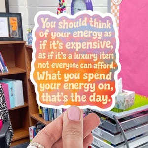 Puede incluir: Una pegatina colorida con el texto "You should think of your energy as if it's expensive, as if it's a luxury item not everyone can afford. What you spend your energy on, that's the day." La pegatina está sujeta por una mano.
