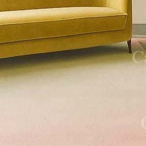 Pu&ograve; includere: Un tappeto dai colori pastello con un effetto sfumato, che passa dal rosa chiaro al bianco crema. Un divano giallo &egrave; parzialmente visibile sullo sfondo. Il tappeto ha un bordo con strisce rosa e verdi.