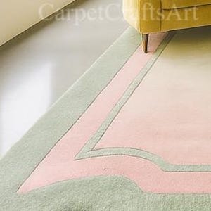 Pu&ograve; includere: Un tappeto dai colori pastello con un design geometrico. Il tappeto presenta un bordo verde chiaro, una sezione centrale rosa e un interno color crema. Il tappeto &egrave; parzialmente visibile, con una sezione di una gamba del divano gialla nell'angolo in alto a destra.