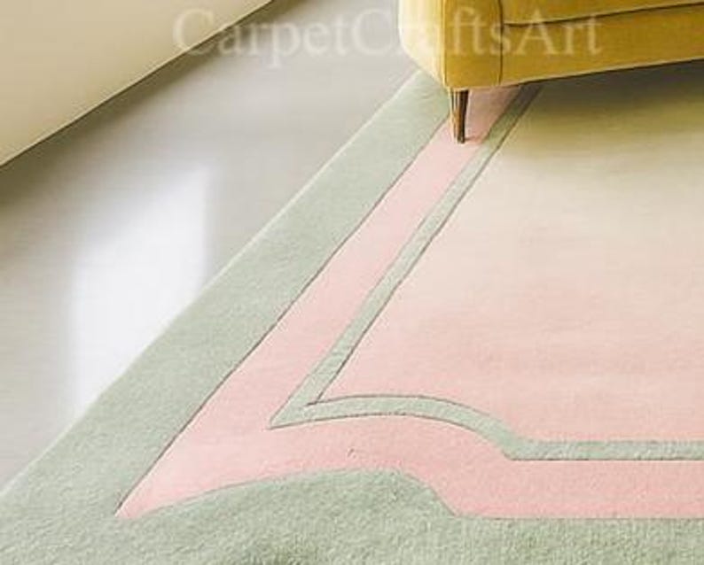 Pu&ograve; includere: Un tappeto dai colori pastello con un design geometrico. Il tappeto presenta un bordo verde chiaro, una sezione centrale rosa e un interno color crema. Il tappeto &egrave; parzialmente visibile, con una sezione di una gamba del divano gialla nell'angolo in alto a destra.