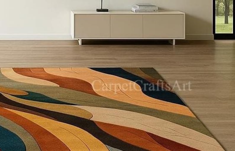 May include: A rectangular rug with an abstract design in shades of orange, brown, teal, and beige. The rug has a wave-like pattern and is on a wooden floor. The text "CarpetCraftsArt" is visible on the rug. The rug is suitable for a living room.