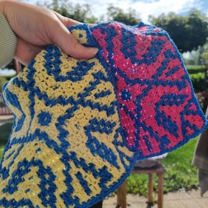 Op de afbeelding: Twee gehaakte vierkanten met geometrische patronen. Eén is geel en blauw, de andere roze en blauw. De vierkanten worden omhoog gehouden tegen een achtergrond van blauwe lucht en groene bomen.