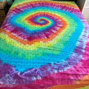 May include: A bed sheet featuring a vibrant tie-dye spiral pattern in shades of red, orange, yellow, green, blue, and purple. The fabric displays a repeating subtle geometric print layered beneath the colorful, swirling tie-dye design.