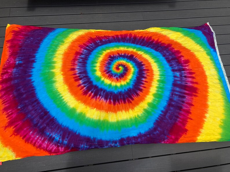 Pu&ograve; includere: Un arazzo tie-dye colorato con un grande motivo a spirale in tonalit&agrave; di rosso, arancione, giallo, verde, blu e viola.