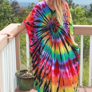 May include: A vibrant tie-dye caftan dress in a spiral pattern of black, red, orange, yellow, green, and blue. The dress has a V-neck and wide sleeves. The person is standing on a wooden deck.