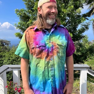 May include: A short-sleeved button-down shirt featuring a vibrant tie-dye pattern in shades of purple, blue, green, orange, and pink. The shirt includes two chest pockets with button closures and a full-length front button placket.