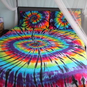 May include: A bed with a tie-dye comforter and matching pillowcases. The comforter has a large, colorful swirl pattern in shades of blue, green, yellow, orange, red, and purple. The bed is topped with a white mosquito net.
