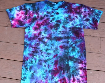 May include: A tie-dye t-shirt in a galaxy-inspired design. The shirt features a blend of blue, purple, and black hues, with white speckles. The short-sleeved shirt is laid flat on a wooden surface.