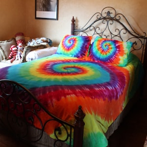 Pu&ograve; includere: Un letto con una struttura in metallo nero e un piumino tie-dye in tonalit&agrave; di blu, verde, arancione, giallo e rosso. Il piumino ha un motivo a spirale.