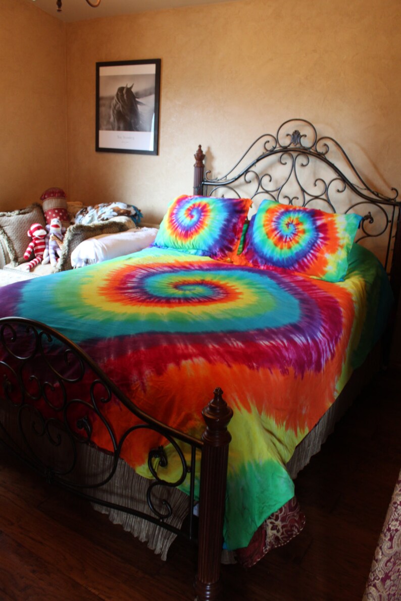 Pu&ograve; includere: Un letto con una struttura in metallo nero e un piumino tie-dye in tonalit&agrave; di blu, verde, arancione, giallo e rosso. Il piumino ha un motivo a spirale.