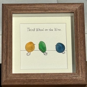 Peut inclure: Une œuvre encadrée avec trois illustrations d'oiseaux colorés sur un fil. Les oiseaux sont jaunes, verts et bleus, avec des pattes simples dessinées au trait. Le texte "Third Wheel on the Wire" est au-dessus des oiseaux.