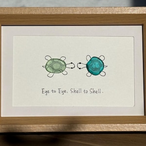 May include: A framed watercolour print with two turtles, one green and one teal, linked by a line. The text reads "Eye to Eye, Shell to Shell". The frame is made of light brown wood.