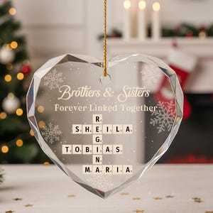 May include: A clear, heart-shaped crystal ornament inscribed with "Brothers & Sisters Forever Linked Together" and names in Scrabble tiles. The ornament hangs from a gold string. A Christmas tree and stockings are visible in the background.