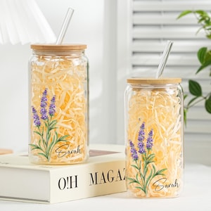 May include: Two clear glass tumblers with bamboo lids and glass straws. Each tumbler has a lavender floral design and the name "Sarah". The tumblers are filled with yellow paper and are on a book.