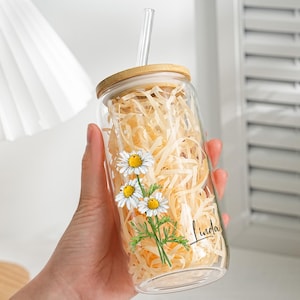 Puede incluir: Un vaso de vidrio transparente con tapa de bambú y pajita transparente. El vaso está decorado con un diseño floral de margaritas y el nombre "Linda" en cursiva. El vaso está lleno de papel triturado.