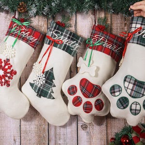 Puede incluir: Cuatro calcetines navideños blancos con nombres bordados en los puños a cuadros. Cada calcetín presenta diseños festivos: un copo de nieve, un árbol de Navidad, un hueso y una huella de pata. Los acentos rojos y verdes realzan el tema navideño.