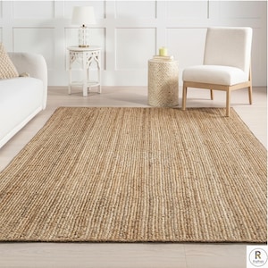 May include: A rectangular jute rug with a natural, light brown color. The rug has a ribbed texture and is placed on a light-colored wooden floor. The rug is in a living room setting, with a white sofa, side table, and chair visible.