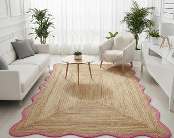 May include: A rectangular jute rug with a scalloped pink border. The rug has a natural beige color and is placed in a living room setting with white furniture and plants. The rug's dimensions are not visible.