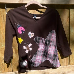Op de afbeelding: Een bruin shirt met lange mouwen en een V-hals, versierd met stoffen applicaties. Applicaties zijn onder meer een lieveheersbeestje, hart, ster, vlinder en appel. Een grote roze, zwart-wit geruite patch bevindt zich aan de rechterkant. Het shirt hangt aan een houten hanger.