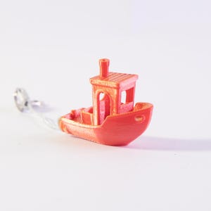 Op de afbeelding: Een kleine, feloranje 3D-geprinte speelgoedboot met een hut en schoorsteen. De boot is verbonden met een zilverkleurig object door een dunne, transparante draad. De boot is een miniatuurmodel, waarschijnlijk een decoratieartikel of een speelgoed.
