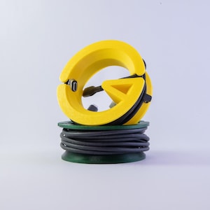 Op de afbeelding: Een geel en zwart kabelbeheergereedschap met een cirkelvormig ontwerp, rustend op een groene basis met een opgerolde zwarte kabel. De tool heeft een driehoekige vorm en een clip.