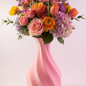 Op de afbeelding: Een kleurrijk boeket bloemen in een roze vaas. Het arrangement bevat oranje tulpen, roze rozen, gele ranonkels en paarse hortensia's. De vaas heeft een wervelend ontwerp.