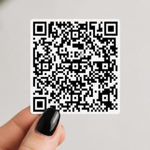 May include: A square, black and white QR code sticker is held up against a white background. The sticker is held by a hand with black nail polish. The QR code is a pattern of black and white squares.