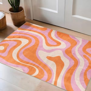 Puede incluir: Una alfombra rectangular con un diseño retro. La alfombra presenta un patrón de líneas onduladas en tonos naranja, rosa y crema. La alfombra está colocada sobre un suelo de madera clara cerca de una puerta blanca.