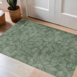 Puede incluir: Una alfombra rectangular verde salvia con un patrón repetido de contornos de hojas blancas. La alfombra está colocada frente a una puerta blanca y tiene un borde claro.