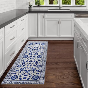 Op de afbeelding: Een rechthoekig keukenkleed met een bloemenpatroon. Het kleed heeft een crèmekleurige achtergrond met een blauw bloemenpatroon en een blauwe Griekse sleutelrand. Het kleed ligt op een donkere houten vloer in een keuken.