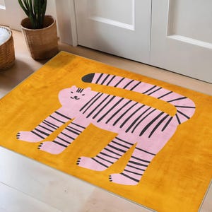 May include: A rectangular rug with a pink tiger design on a yellow background. The tiger has black stripes and is in a walking pose. The rug is placed on a light-colored wooden floor.