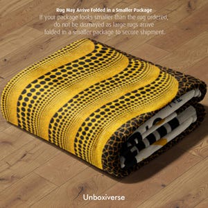 Op de afbeelding: Gevouwen tapijt met een geel en zwart patroon. Het tapijt heeft een patroon van zwarte stippen en strepen. Het lijkt gemaakt van een zacht materiaal en is opgevouwen. De tekst "Rug May Arrive Folded in a Smaller Package" is zichtbaar.
