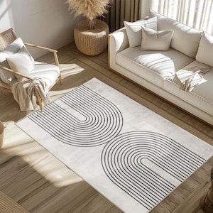 Può includere: Un tappeto rettangolare con sfondo crema e due grandi disegni ad arco neri simmetrici. Il tappeto è posizionato su un pavimento in legno, con un divano bianco, una sedia ed elementi decorativi sullo sfondo. Il design del tappeto aggiunge un tocco moderno.