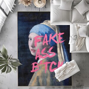 May include: A rectangular rug featuring a portrait of a woman with a blue headscarf and pearl earring, with the words "FAKE ASS BITCH" in pink lettering. The rug has a dark background and is surrounded by furniture.