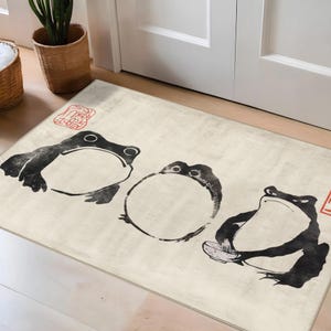 Op de afbeelding: Een rechthoekige beige deurmat met drie gestileerde zwarte kikkerillustraties. De kikkers zijn afgebeeld in een minimalistische stijl, één houdt een kleine kom vast. Rode Japanse karakters in de hoeken.