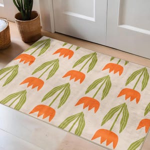 May include: A rectangular beige rug with a repeating pattern of orange tulip-like flowers and green leaves. The rug is placed in front of a white door and has a simple, modern design.