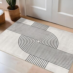 May include: A rectangular rug with a geometric design. The rug features a cream-colored background with a black linear pattern. The pattern consists of straight and curved lines that intersect to create an abstract design. The rug is placed on a wooden floor.