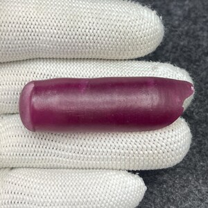 Puede incluir: Una gema pulida y alargada en un tono magenta intenso. La piedra tiene un extremo redondeado y un borde ligeramente irregular y astillado en el extremo opuesto. La gema se muestra sobre una tela blanca texturizada.