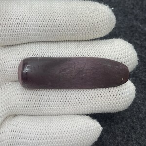May include: A smooth, elongated, purple stone with rounded ends. The stone has a gradient of color, transitioning from a lighter purple to a darker shade. The object is held by a white glove.
