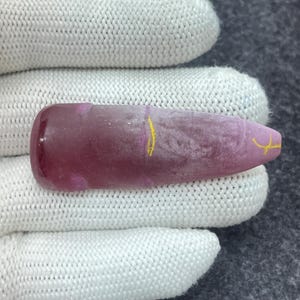 May include: A polished, elongated gemstone with a rounded end and a pointed tip. The stone is a deep, translucent pink with a few yellow markings. The gemstone is held by a person wearing a white glove.
