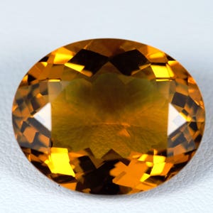 May include: An oval-cut citrine gemstone with a deep golden-yellow hue and a faceted surface. The stone displays a high level of clarity and brilliance, with numerous geometric facets reflecting light across its polished, transparent surface.
