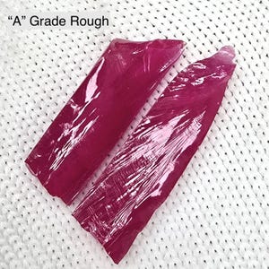 May include: Two rough, deep magenta-colored gemstone pieces, wrapped in clear plastic, are displayed on a white, textured surface. The text "A Grade Rough" is visible in the upper left corner. The gemstones have a raw, natural appearance.